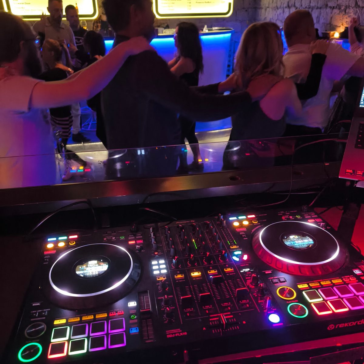 Tanzende Gäste bilden eine Kette vor der beleuchteten Bar, im Vordergrund der DJ-Controller